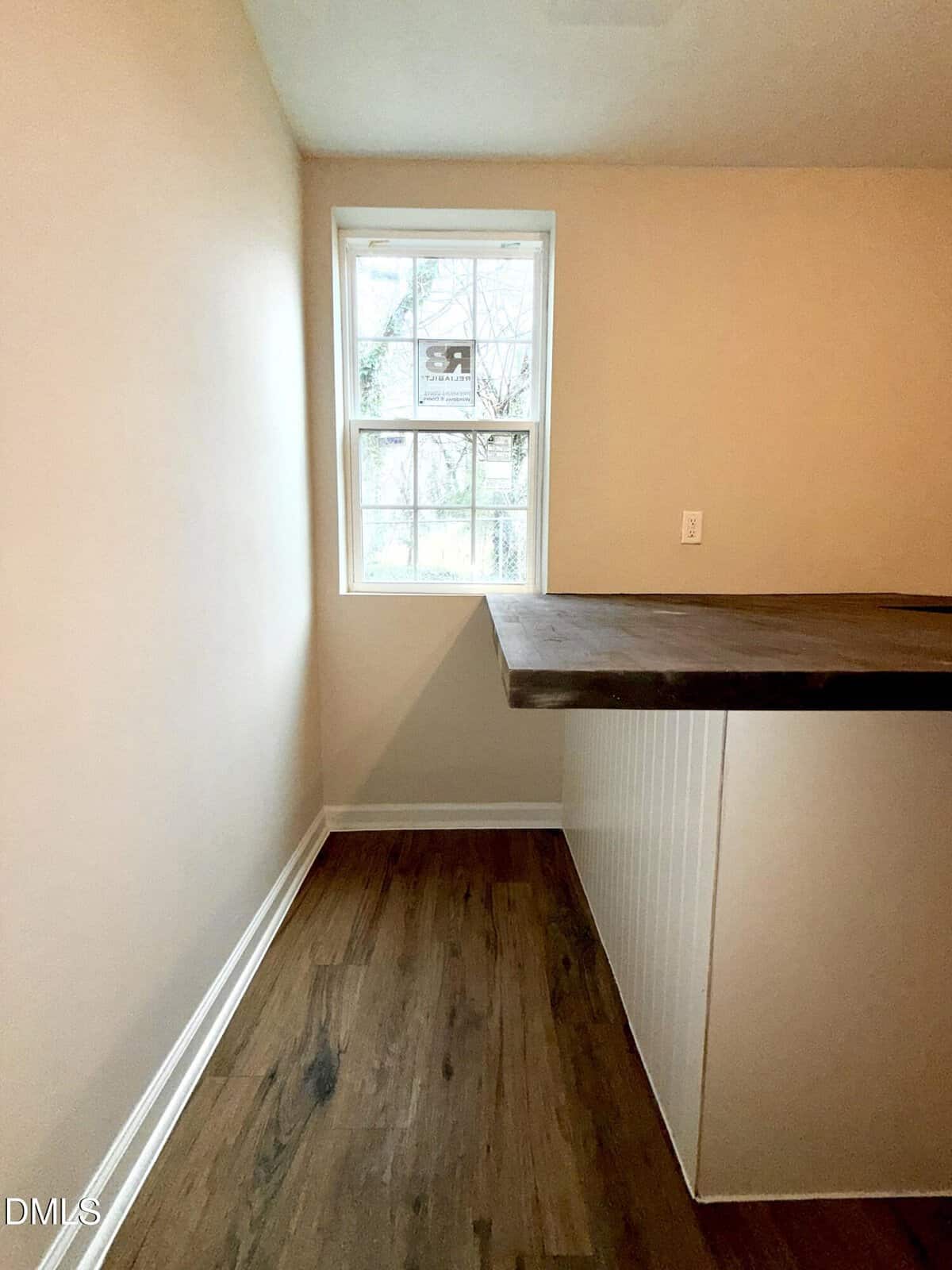 A small, empty room with light-colored walls, a wooden floor, a single window, and a counter with a dark wooden top extending from the right side. Natural light is coming through the window.