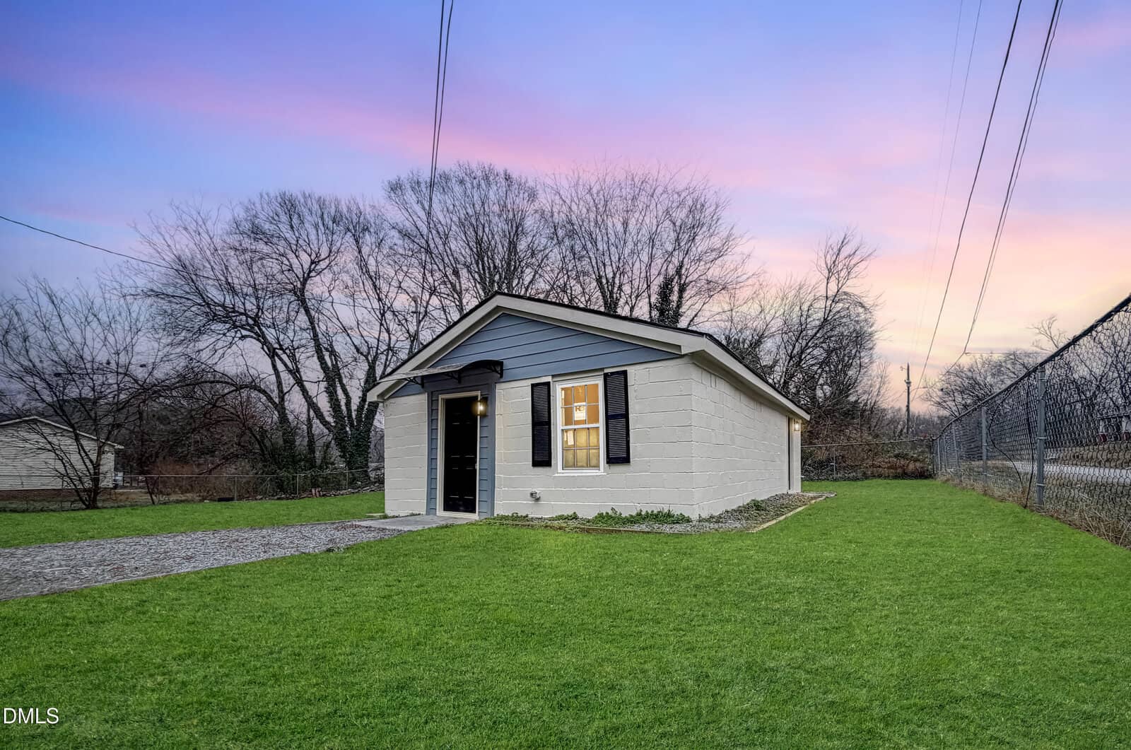 A small, single-story white building with a gray roof sits on a grassy lawn at sunset at 636 Flint Street in Henderson. The building features a black door and window shutters, with bare trees and a chain-link fence in the background.