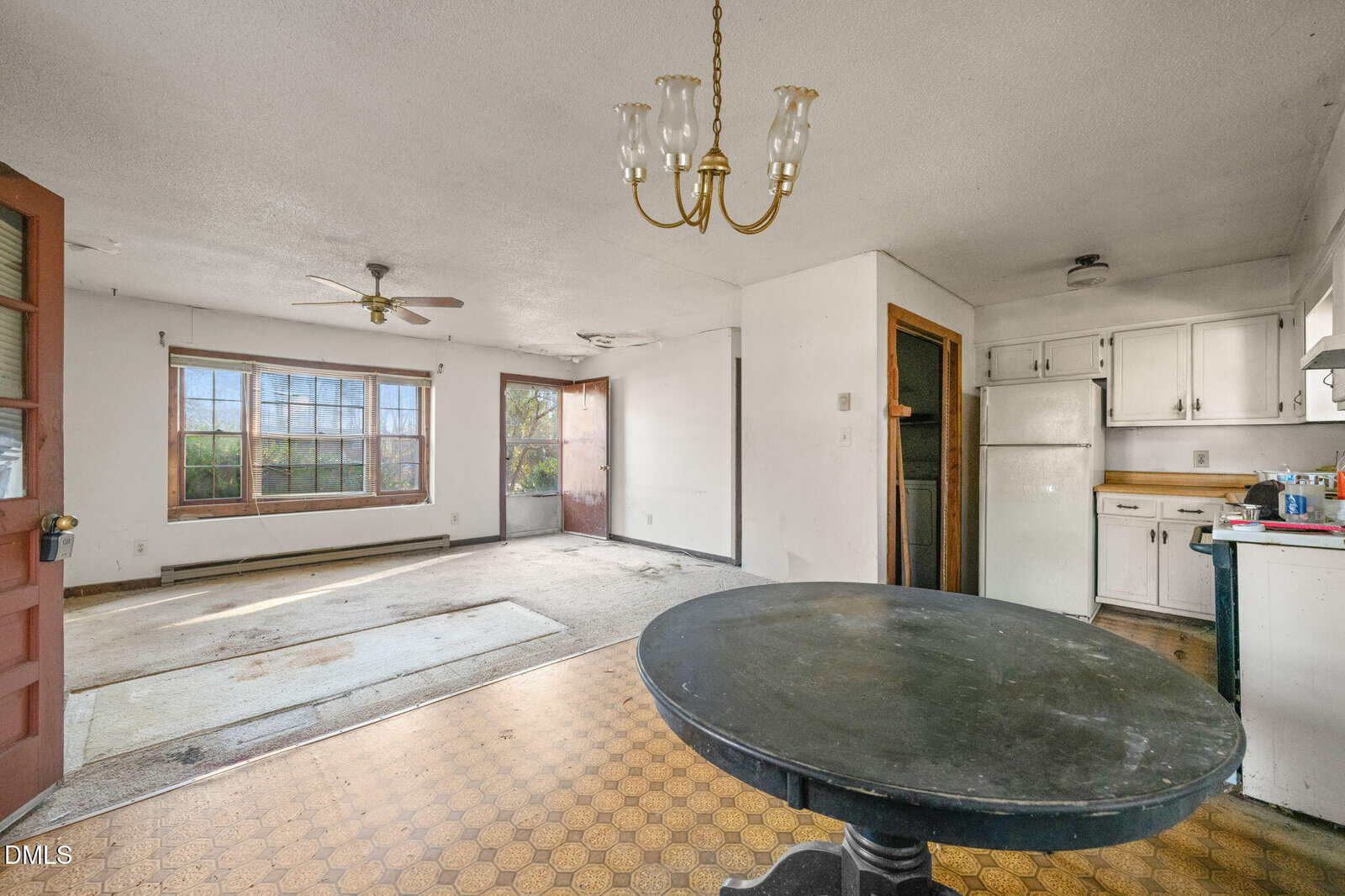 Open kitchen and dining area with old flooring, white cabinets, a round black table, ceiling fans, and large windows letting in natural light. The room appears dated and partially empty.