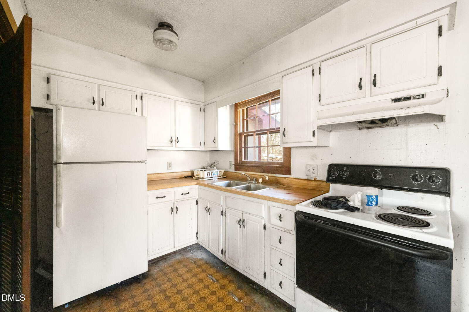 Small, dated kitchen with white cabinets, a white refrigerator, black stove, stainless steel sink under a window, and worn hexagonal-patterned linoleum floor. Some cabinet doors appear slightly damaged or uneven.