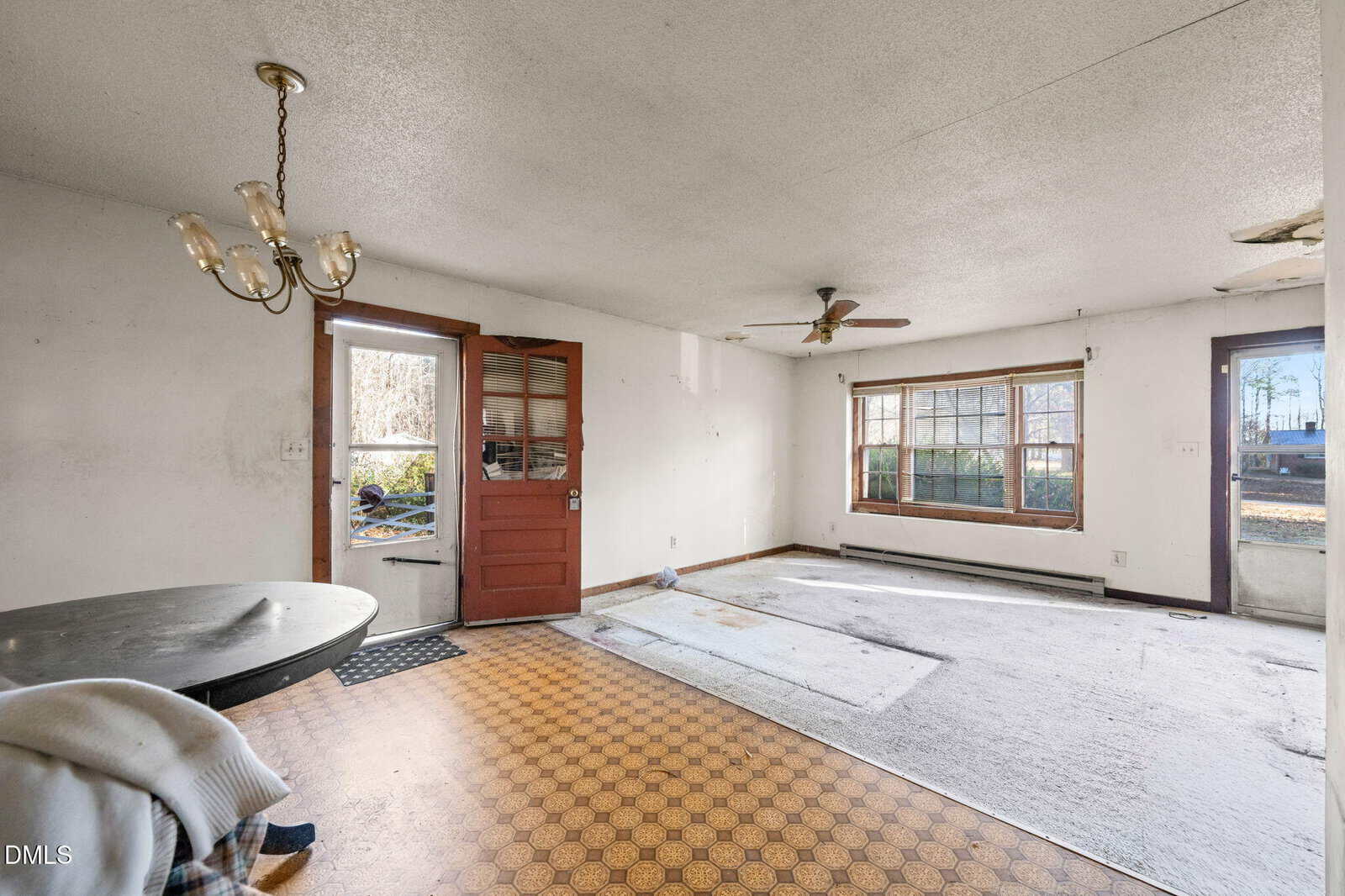 A worn living space with stained carpet, peeling ceiling, gold chandelier, round table with chair and blanket, red door, large window, and visible yard outside. The floor changes from linoleum to carpet.