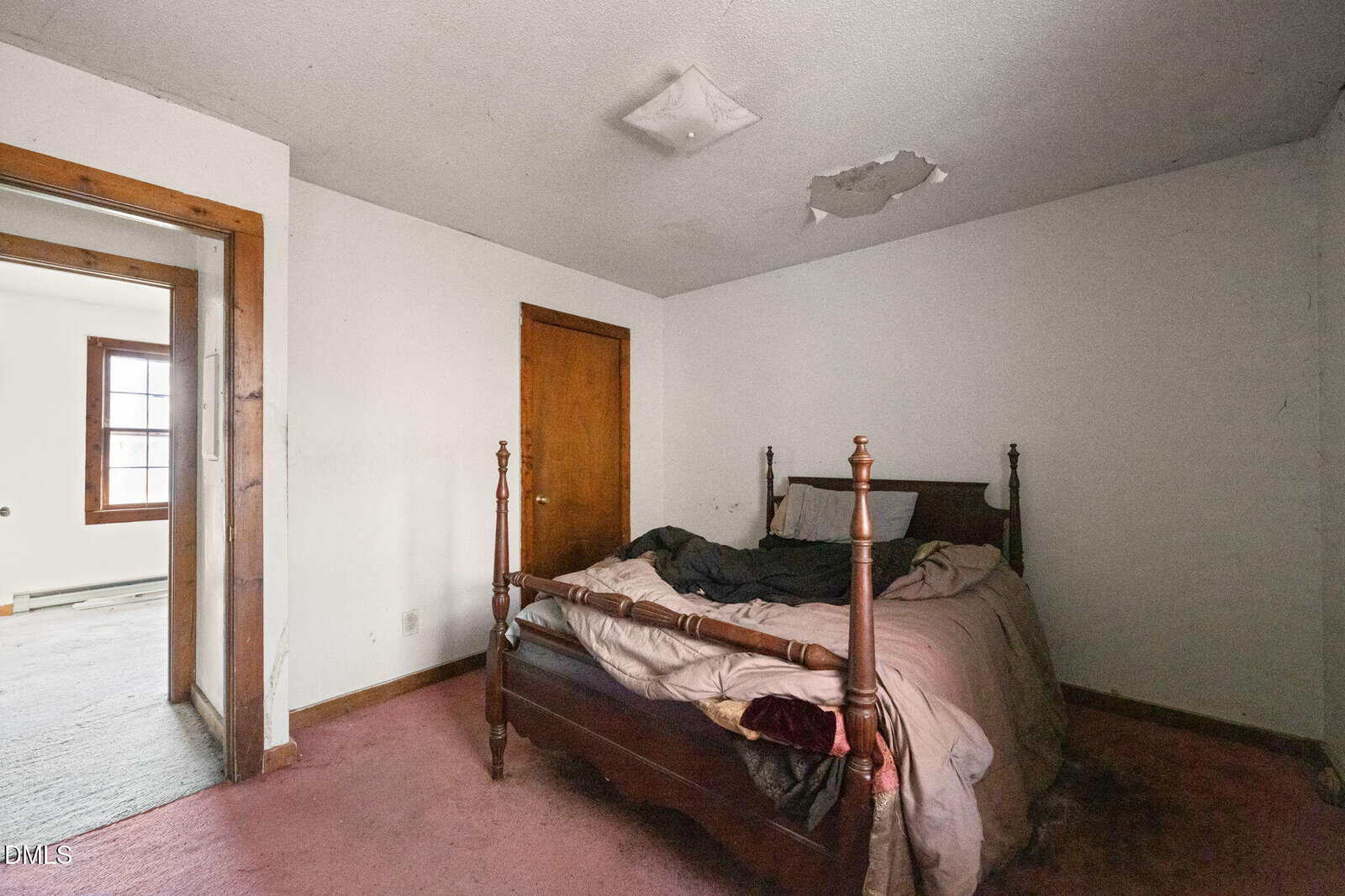 A small, sparsely furnished bedroom with stained carpet, a wooden bed with rumpled bedding, peeling paint on the ceiling, and a door leading to an adjacent room with a window.