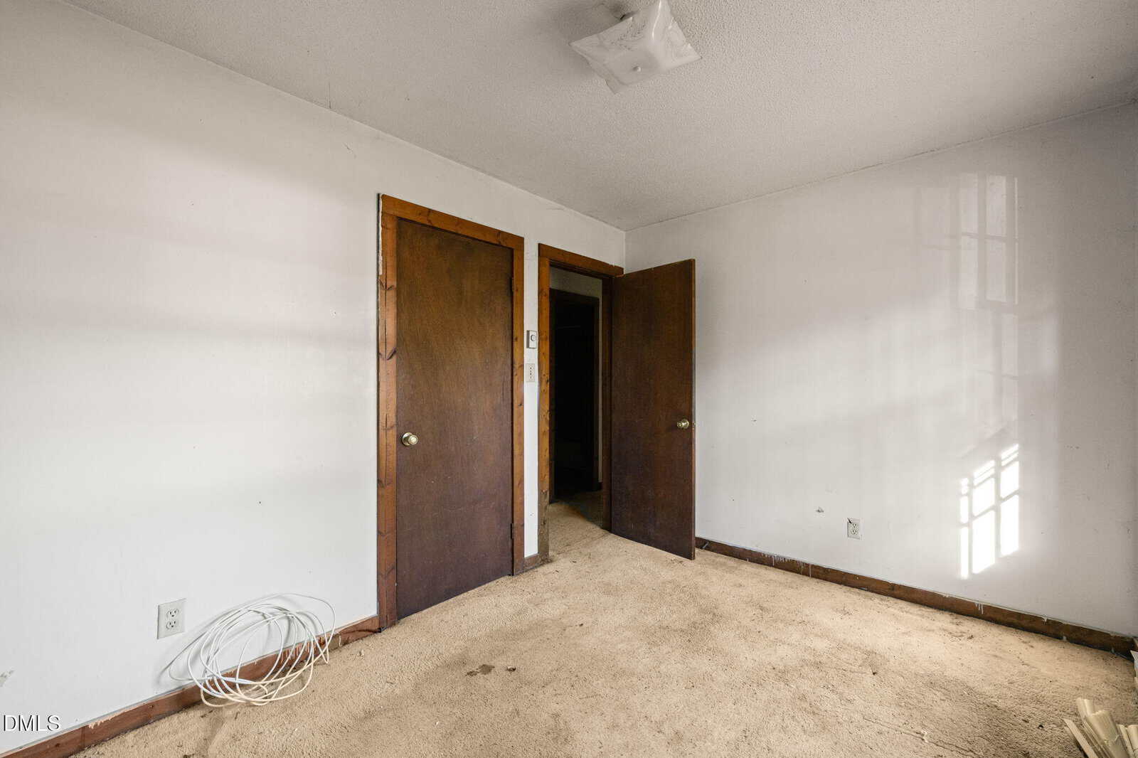 An empty room with beige carpet, white walls, two wooden doors, a ceiling light fixture, and a coiled cable on the floor near an outlet. Sunlight comes through a window, casting shadows on the wall.
