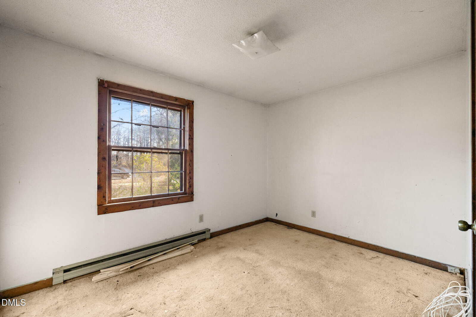 An empty room with white walls, beige carpet, a single window with wooden trim, and a baseboard heater. A few wooden planks lie on the floor beneath the window. The ceiling has a simple light fixture.