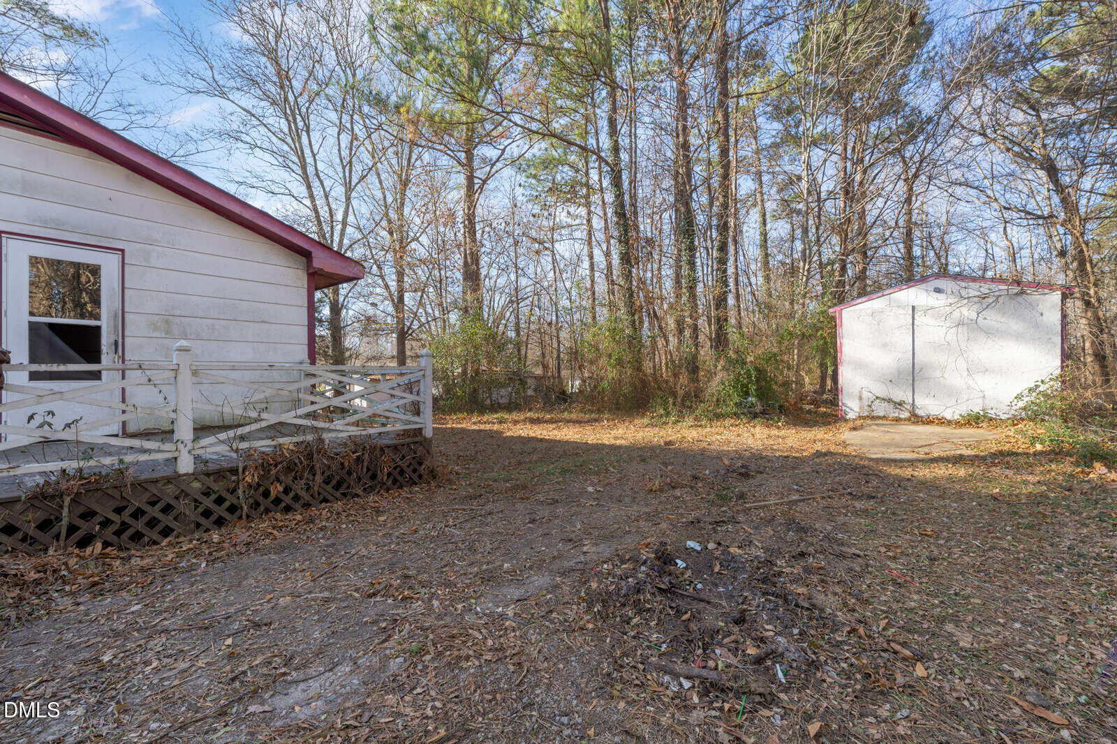 A backyard scene with a small white shed and a house with a deck on the left. The ground is mostly dirt with scattered leaves, and trees without leaves surround the area under a partly cloudy sky.