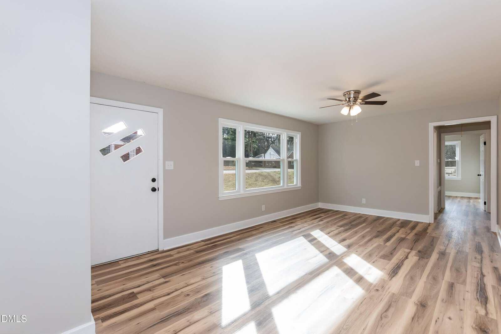 Empty living room with light gray walls, wood-style flooring, a white front door, two windows letting in sunlight, a ceiling fan with lights, and an open doorway leading to another room.