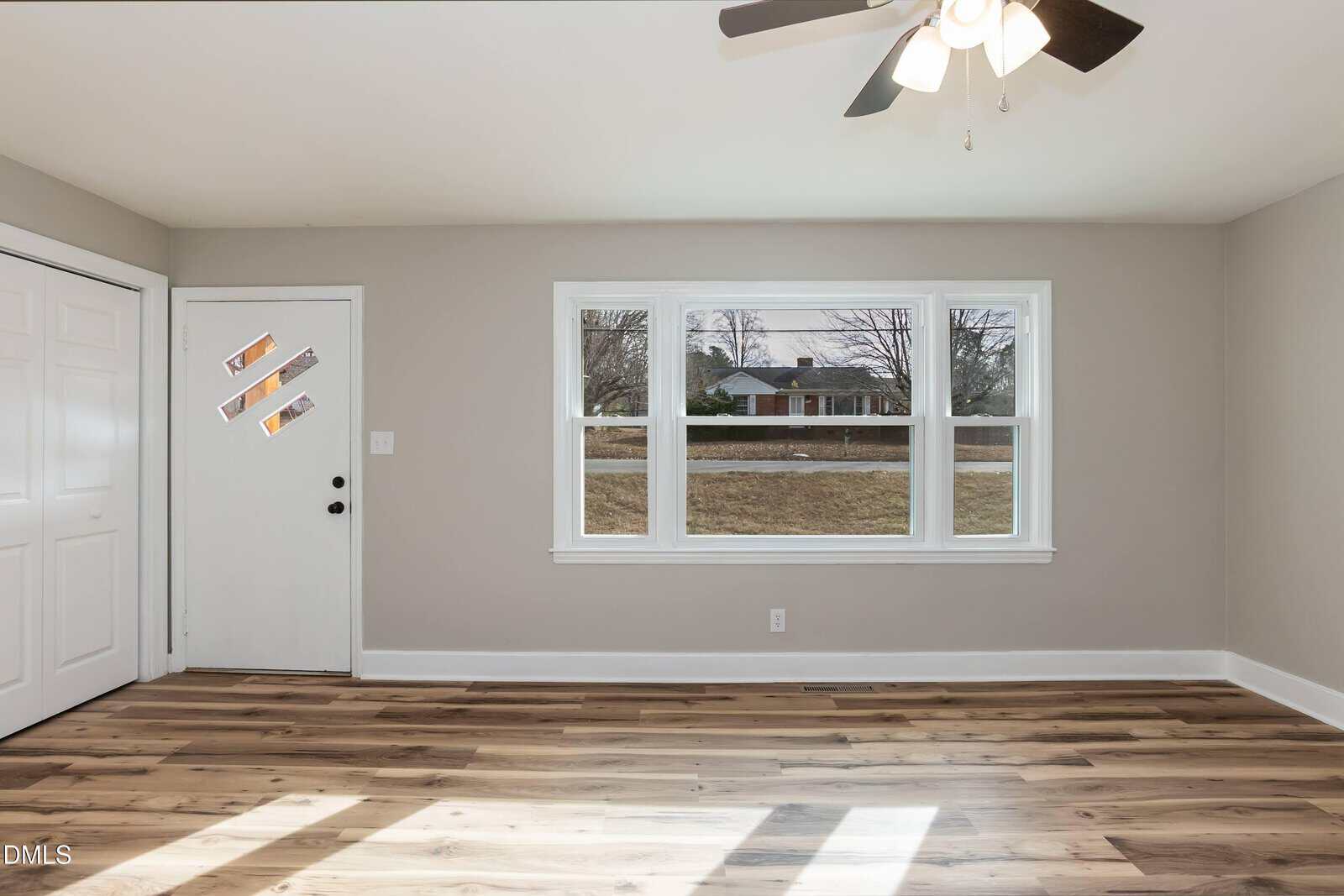 A bright, empty room with light gray walls, wood-look flooring, a ceiling fan with lights, a white door with three diagonal glass panels, double closet doors, and a large window facing the street.