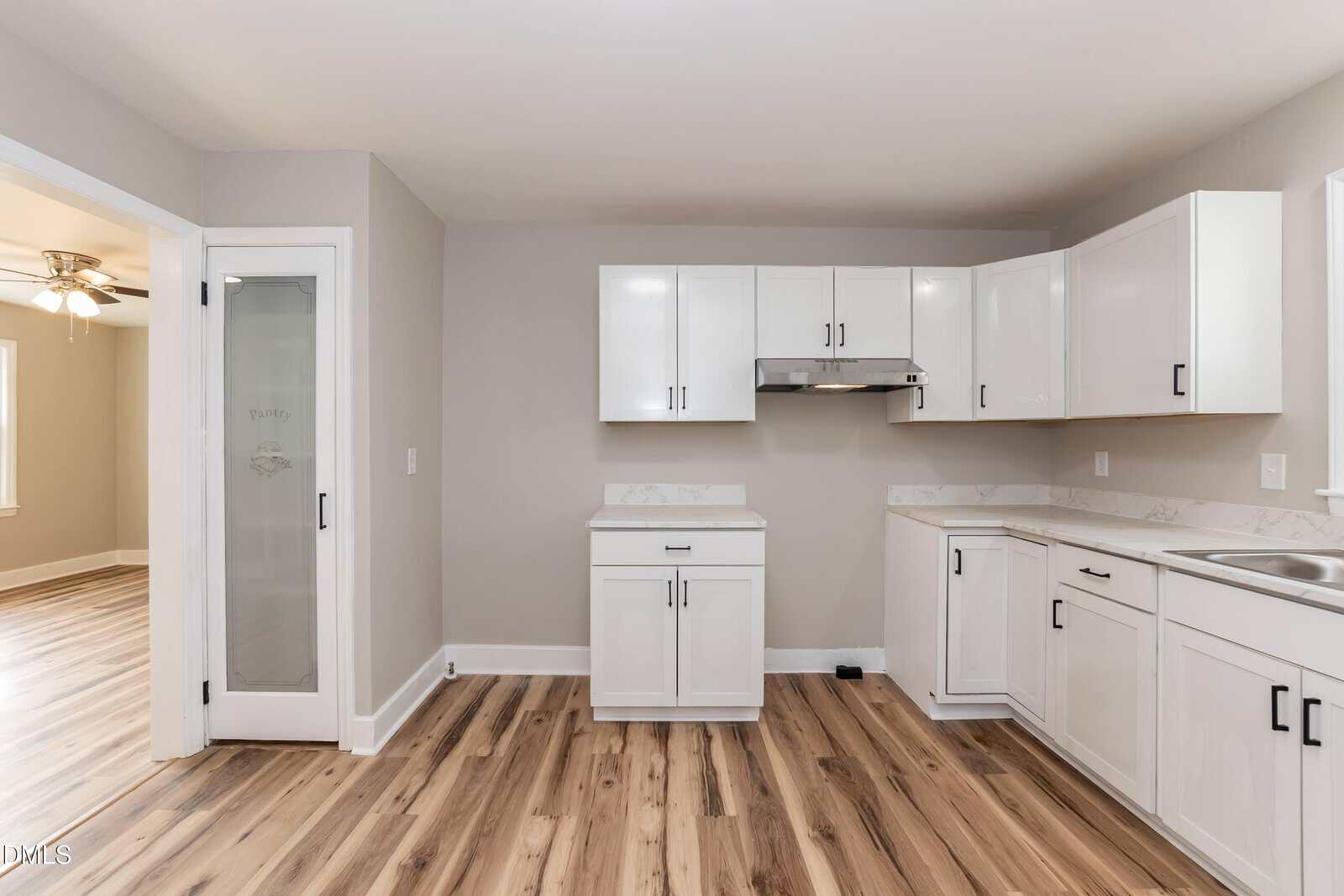 Modern kitchen with white cabinets, light wood flooring, a stainless steel range hood, and a frosted glass pantry door. Space is clean, minimal, and well-lit with neutral wall colors.