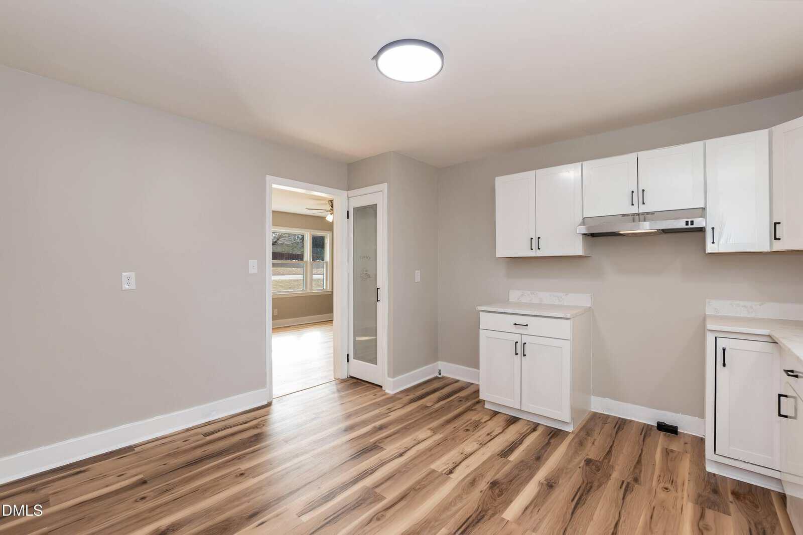 Modern kitchen with white cabinets, light wood flooring, and a glass door. The room is empty, well-lit, and has neutral-colored walls. An adjacent room with large windows is visible through the open doorway.
