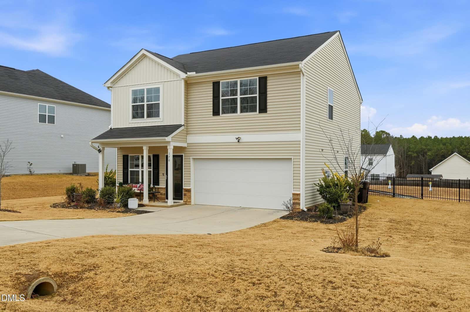 A two-story beige house with black shutters, a covered front porch, and an attached two-car garage sits on a large lot with dry grass and minimal landscaping under a blue sky.