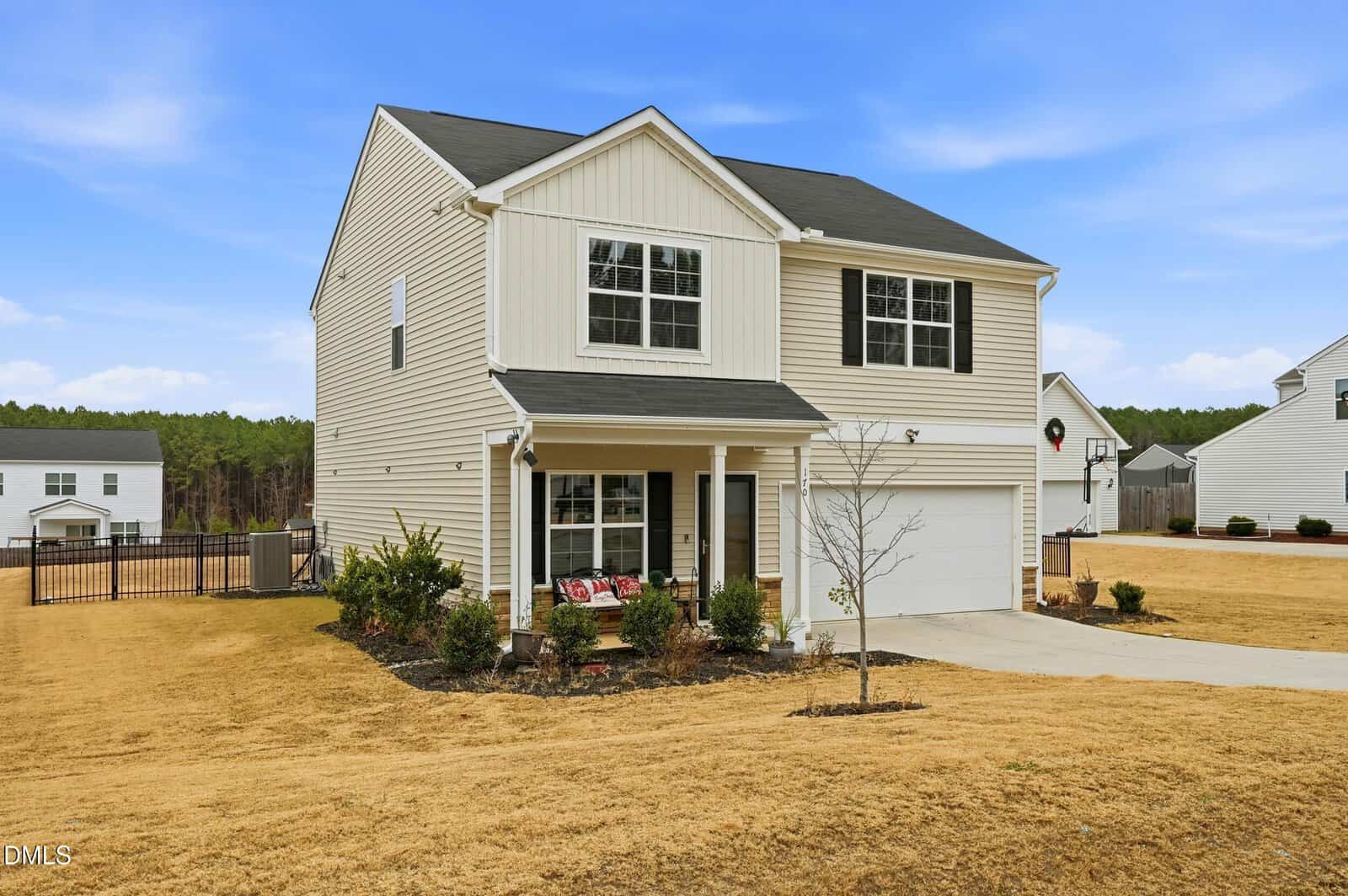 Two-story beige house with white trim and black roof, attached two-car garage, small covered front porch with chairs, minimal landscaping, and a bare tree in the front yard. Surrounding homes and a clear sky are visible.