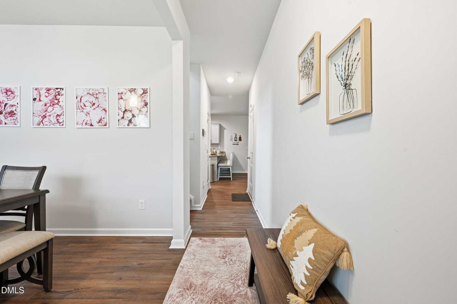 A hallway leads to another room, with a bench, decorative pillow, and pink rug in the foreground. Two framed botanical prints hang above the bench, and pink floral art is visible on the adjacent dining area wall.