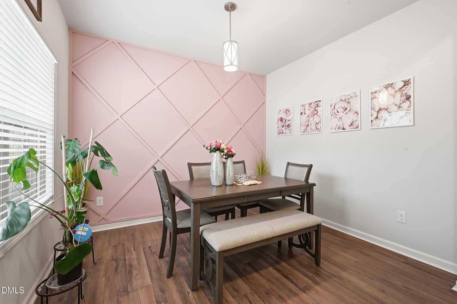 A dining room with a dark wood table, four chairs, and a bench, set on a wood floor. A pink geometric accent wall features three framed artworks. A potted plant and floral centerpiece add decorative touches.