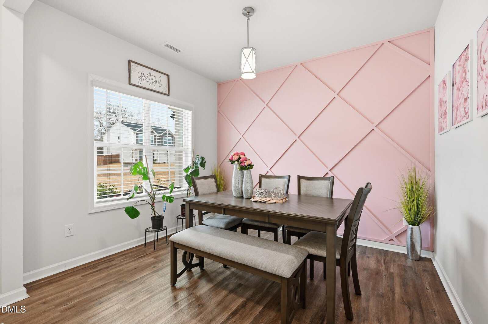 A dining room with a wooden table, six chairs, and a bench, set on hardwood flooring. One wall has pink geometric paneling, and there are plants, wall art, and a window letting in natural light.