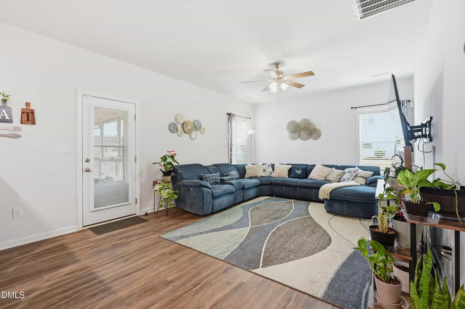 A living room with a large blue sectional sofa, a patterned area rug, several potted plants, wall decor, and a wall-mounted TV. The space features wood flooring and large windows with natural light.