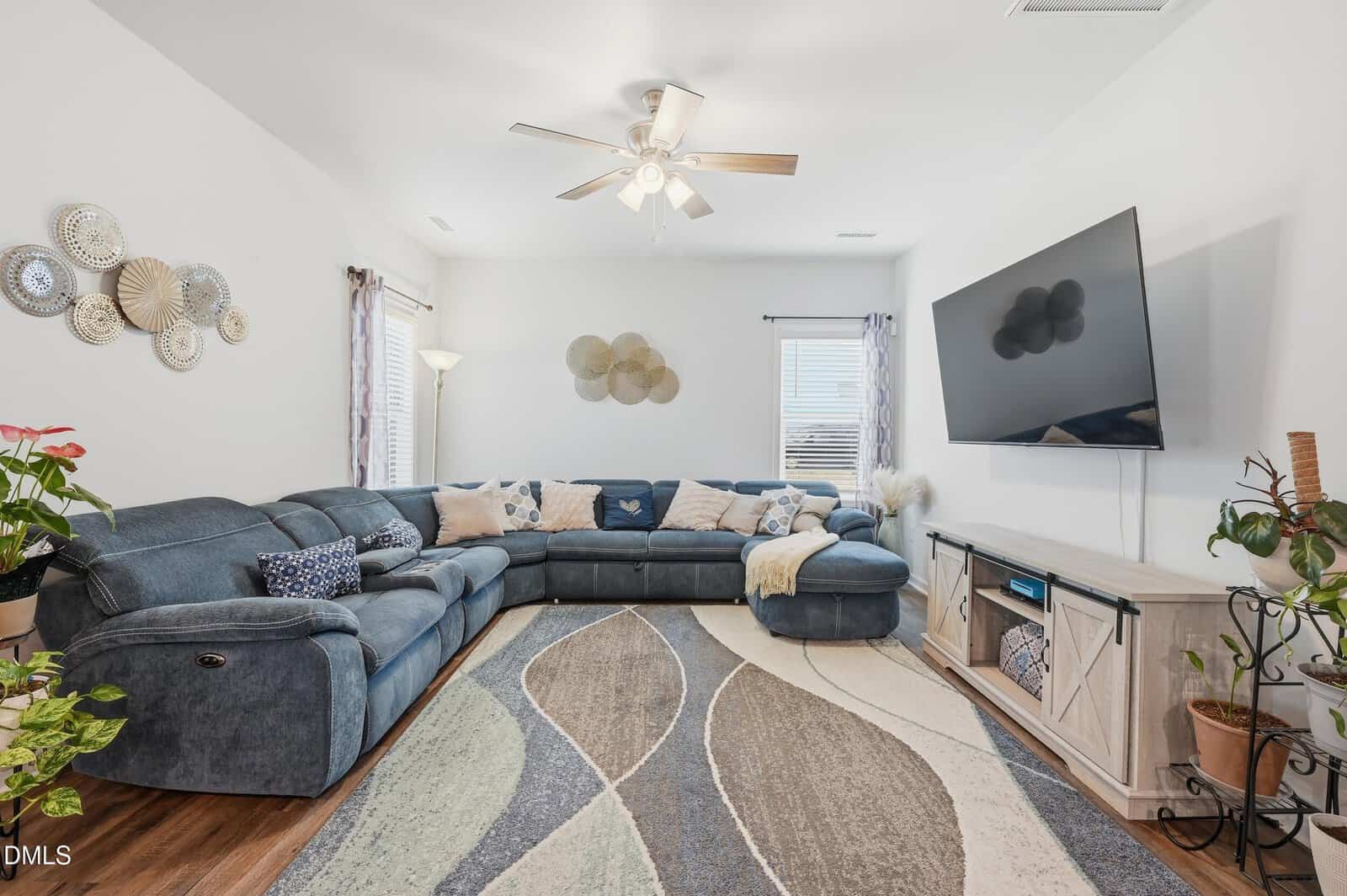 A living room with a large blue sectional sofa, patterned throw pillows, a geometric rug, wall art, a ceiling fan with lights, a wall-mounted flat-screen TV, potted plants, and two windows with sheer curtains.