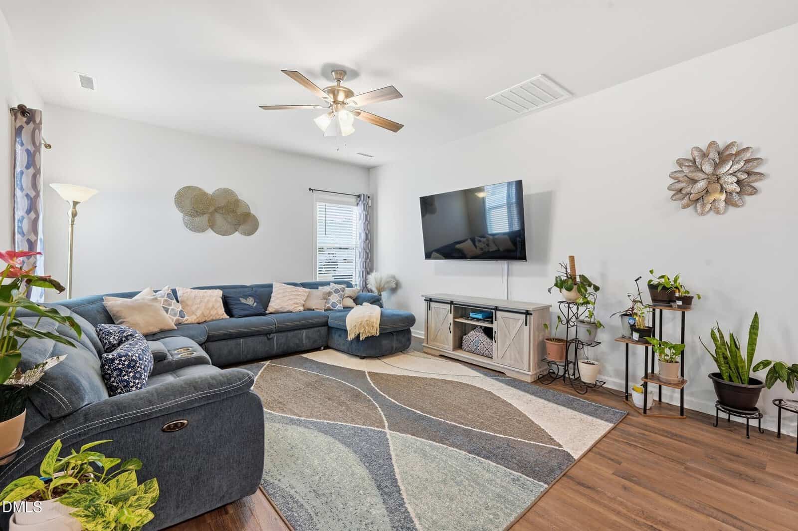 A living room with a large sectional sofa, wall-mounted TV, console table, houseplants, decorative wall art, and a ceiling fan. A geometric rug covers the wood floor and natural light comes through two windows.