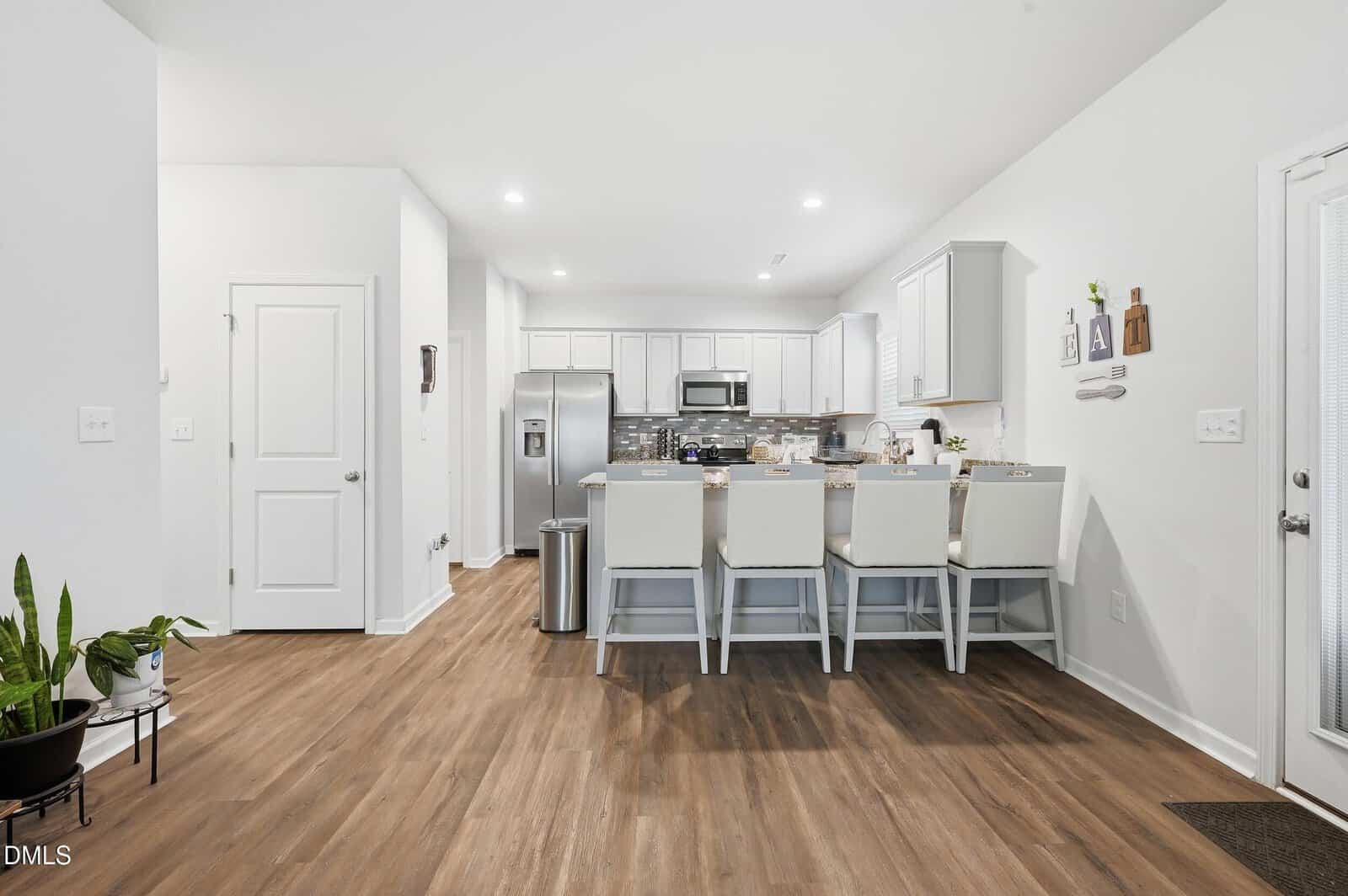 Modern kitchen with white cabinets, stainless steel appliances, and a light wood floor. Four white chairs are lined up at a kitchen island, and a few plants and wall decorations are visible.