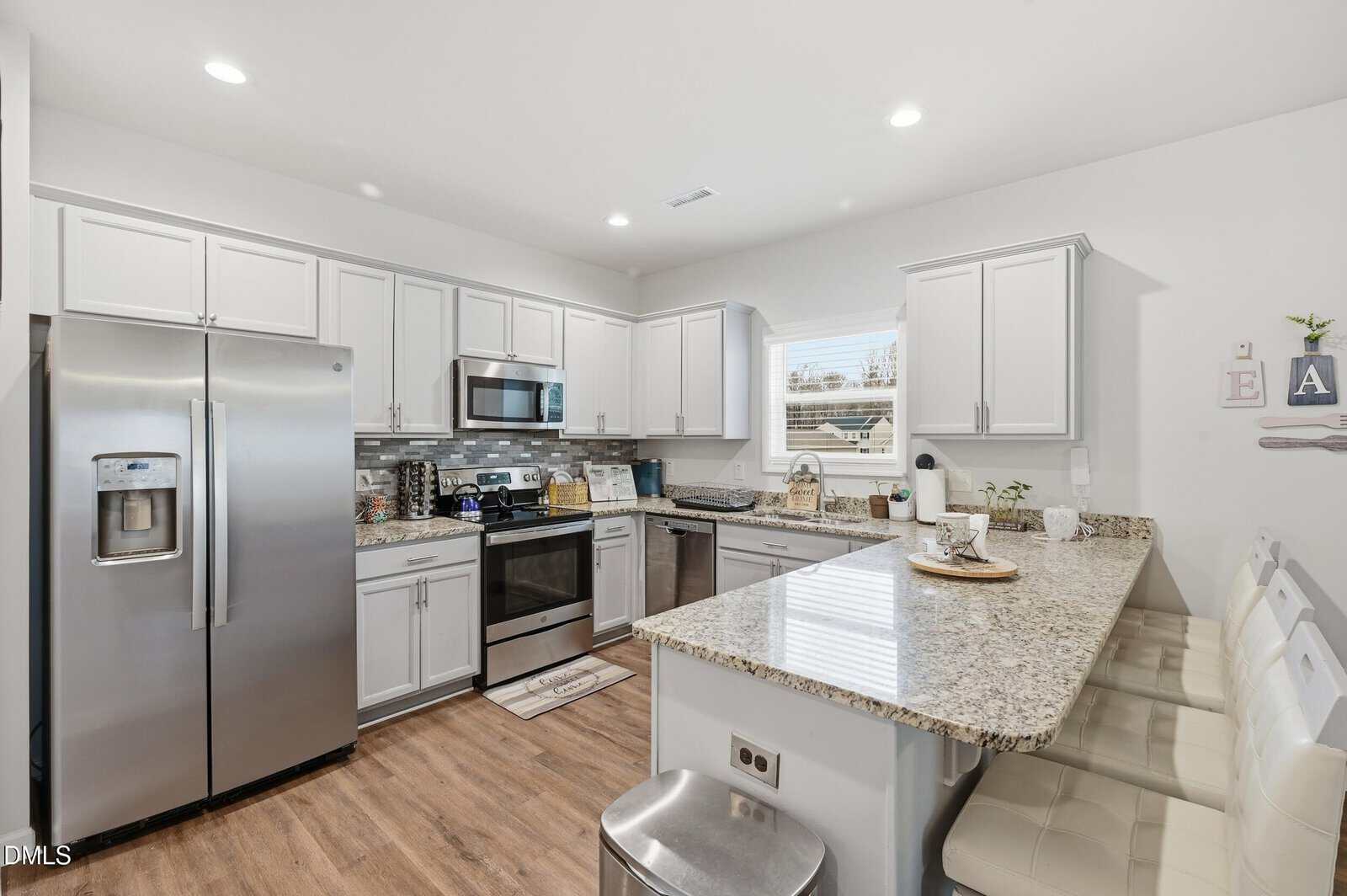 Modern kitchen with stainless steel appliances, white cabinets, granite countertops, and a breakfast bar with three cushioned stools. A window above the sink lets in natural light.