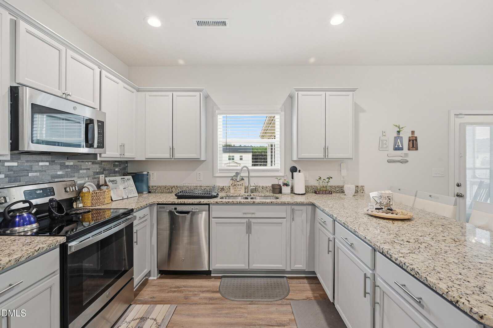 Modern kitchen with white cabinets, stainless steel appliances, granite countertops, and a window above the sink. Decor and kitchen items are neatly arranged on the counters, with light-colored walls and wood flooring.