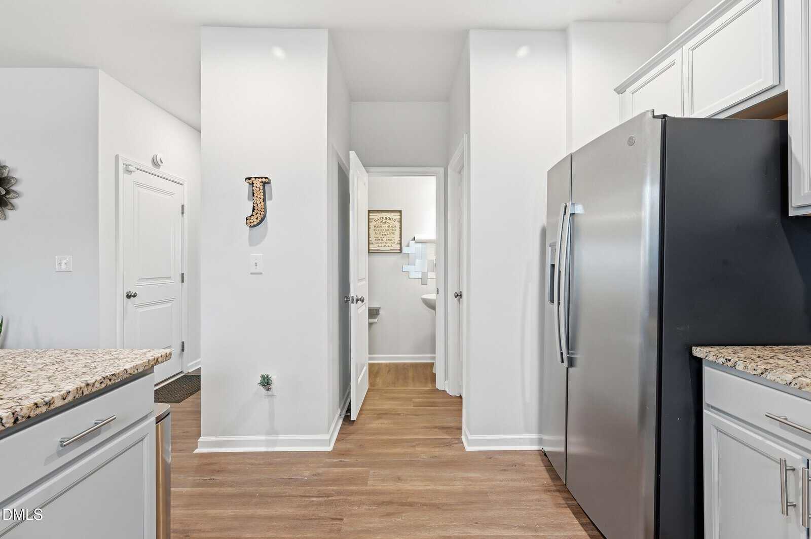 Modern kitchen with light cabinets, granite countertops, stainless steel fridge, and wood floors. Two open doors reveal a bathroom and a room with wall decor. Walls are white, and space is brightly lit.
