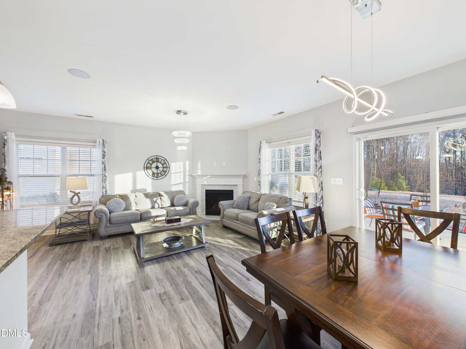 Bright, modern living and dining area with two gray sofas, a fireplace, wood floors, large windows, and a glass dining table. Sliding doors open to a patio with a wooded view.