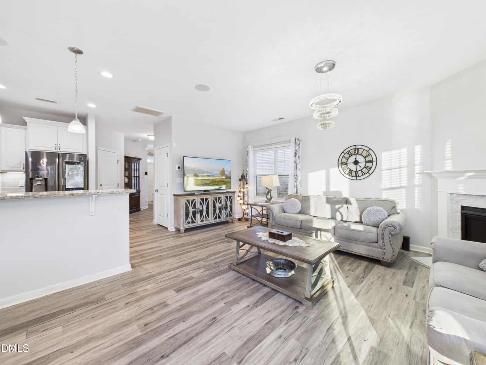 Bright living room with gray sofas, a wooden coffee table, wall clock, TV, and white kitchen with marble countertops. Sunlight streams in through the windows, highlighting the modern, airy decor.