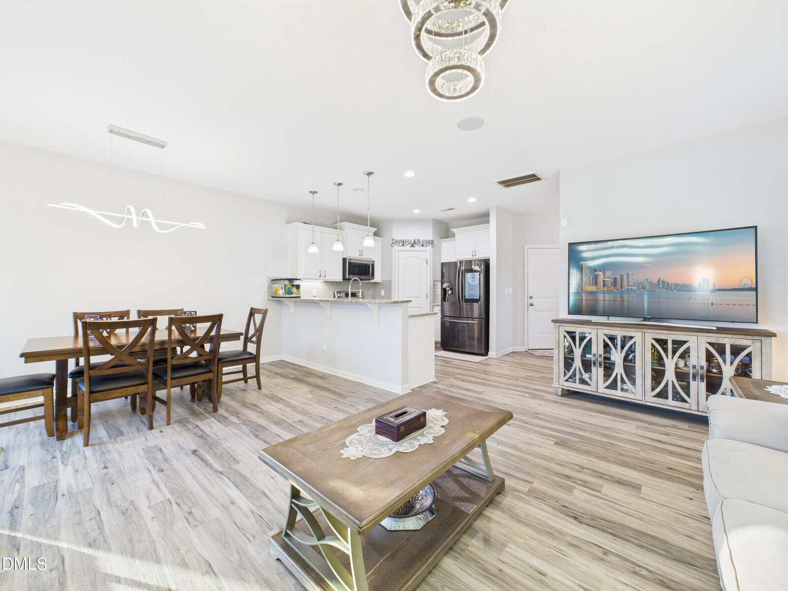 Bright open-concept living area with wood floors, modern chandelier, dining table, kitchen with white cabinets and island, and a large TV on a console.