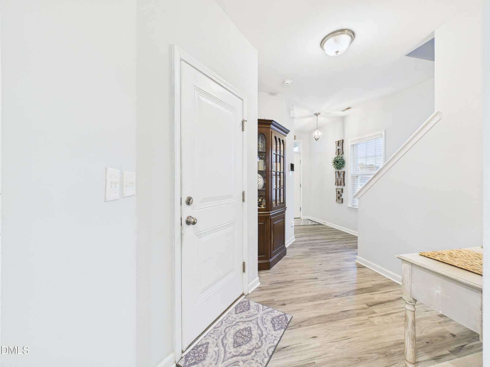 A white front door opens into a hallway with light wood flooring, a patterned rug, a wooden display cabinet, a staircase with white railing, and a window letting in natural light.