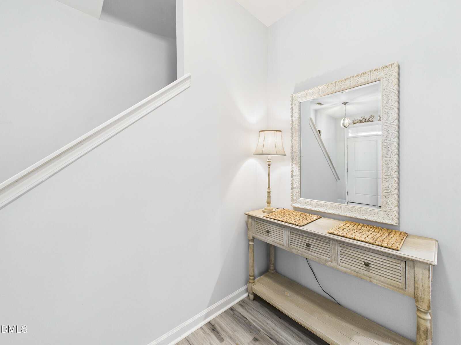 A small entryway with a light wooden console table, two placemats, a decorative lamp, and a large framed mirror. The walls and staircase are painted white, and the floor has light-colored wood planks.
