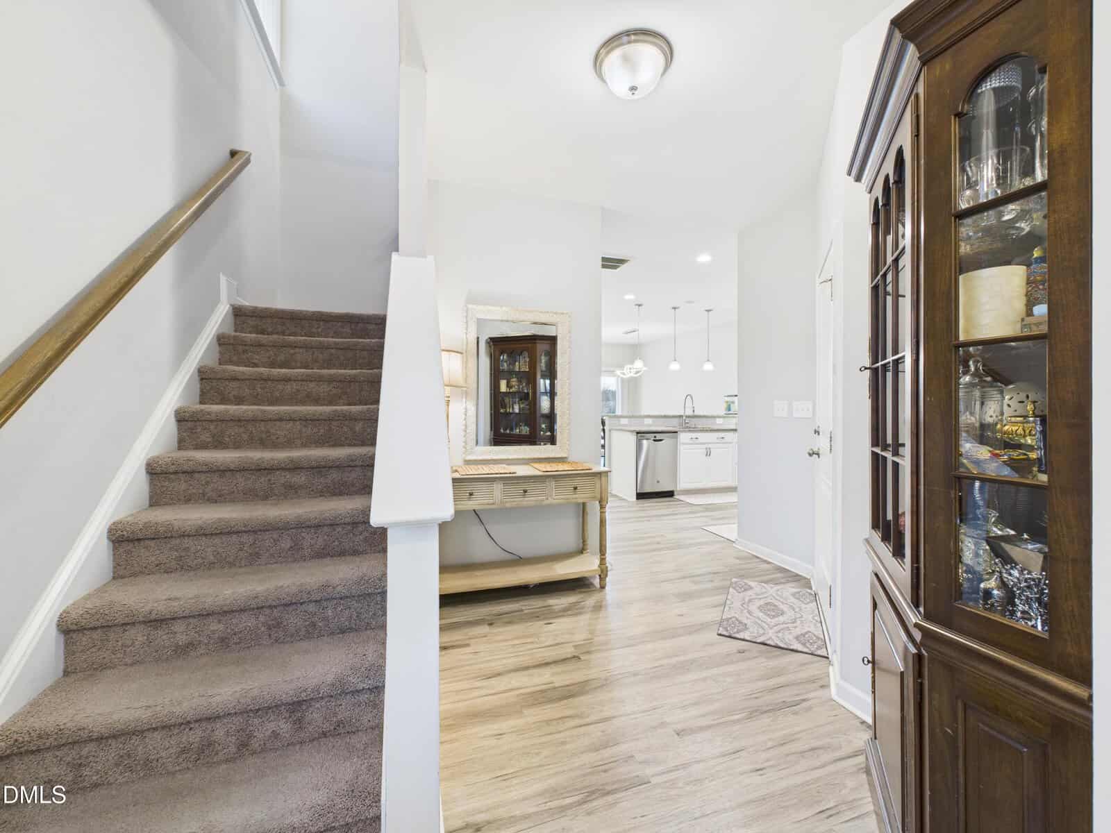 A hallway with a carpeted staircase on the left, a wooden console table with a mirror against the back wall, and a china cabinet on the right. The hallway leads to a kitchen with white cabinets.