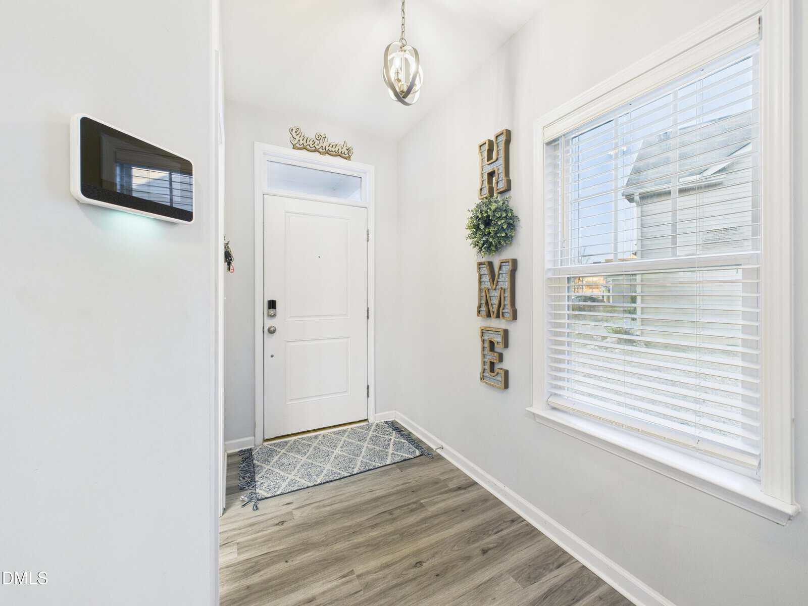 A bright entryway with a white front door, a small mat, wood floors, a wall-mounted control panel, a window with blinds, and decorative "HOME" letters with a wreath hanging vertically on the wall.