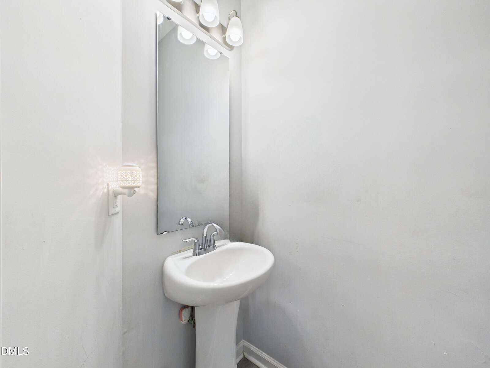 A small, minimalist bathroom with white walls, a pedestal sink, a rectangular mirror, and overhead lighting. There is a sconce on the left wall and visible scuff marks on the walls.