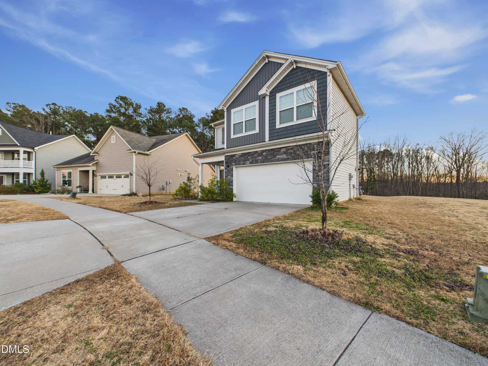 A two-story suburban house with gray siding, white trim, and a stone accent facade. The home has a double garage and driveway, with neighboring houses and a grassy yard under a clear blue sky.