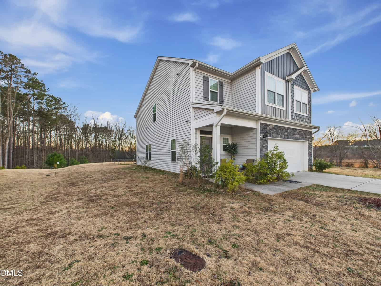 A two-story house with white siding and stone accents sits next to a driveway and attached garage. The lawn is brown and patchy, and the home is surrounded by trees and an open field under a blue sky.