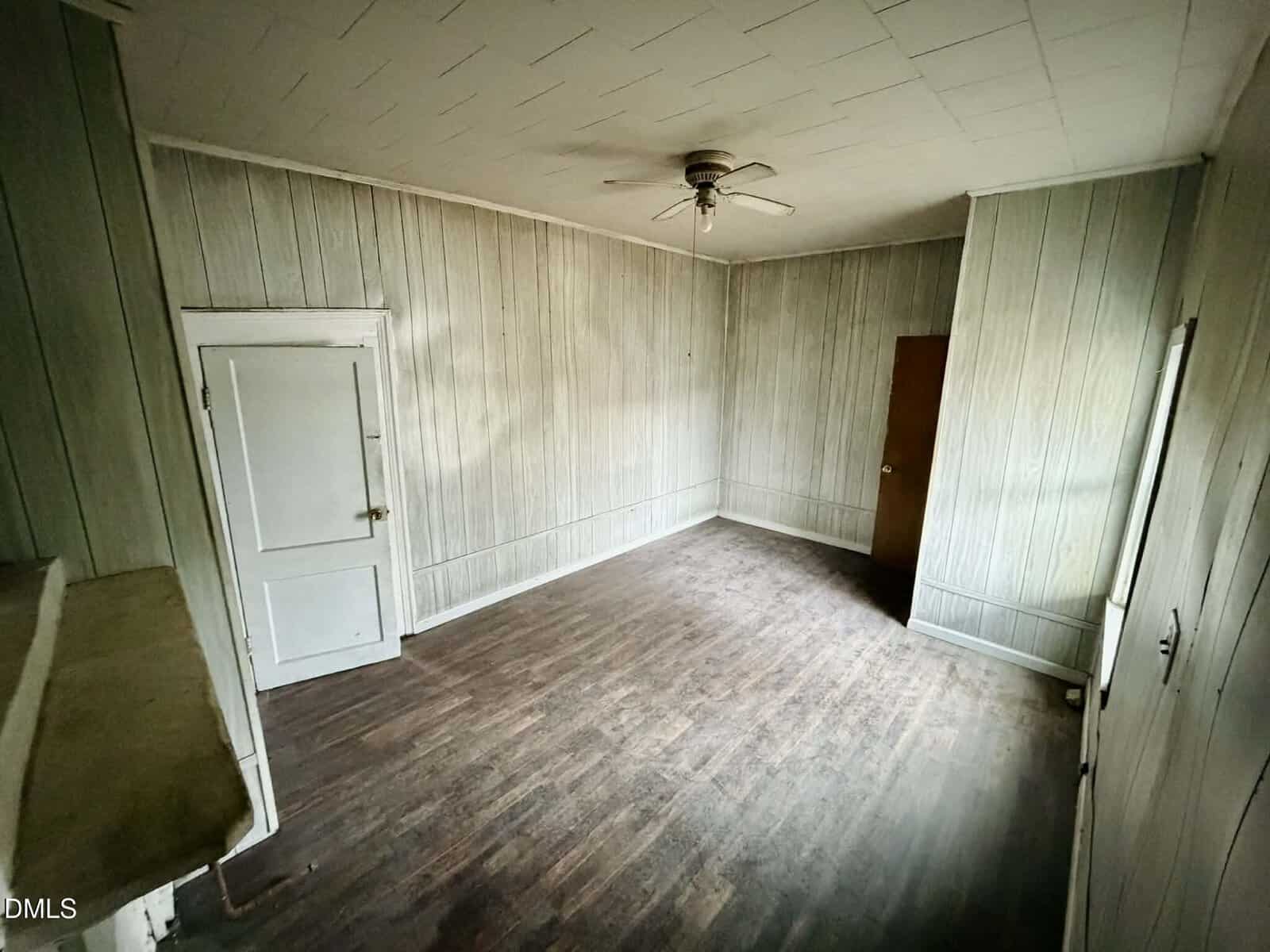 Empty room with wood-paneled walls, a white door, a brown door, ceiling fan, and wood floor. The room appears worn and minimally lit by natural light from a window on the right.