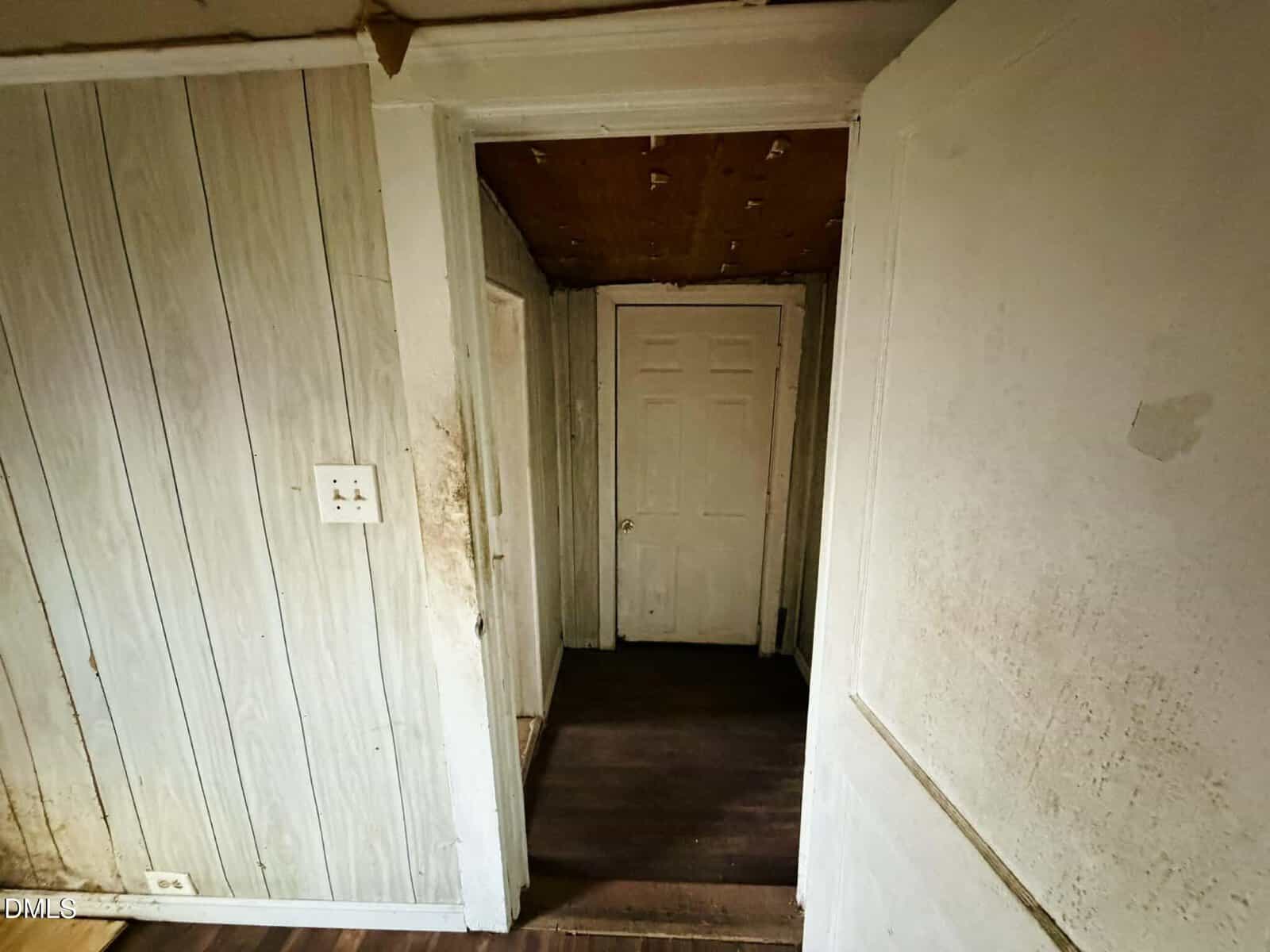 A narrow hallway with worn wood-paneled walls, dirty surfaces, and a closed white door at the end. The ceiling shows signs of damage, and the area appears dimly lit and in poor condition.