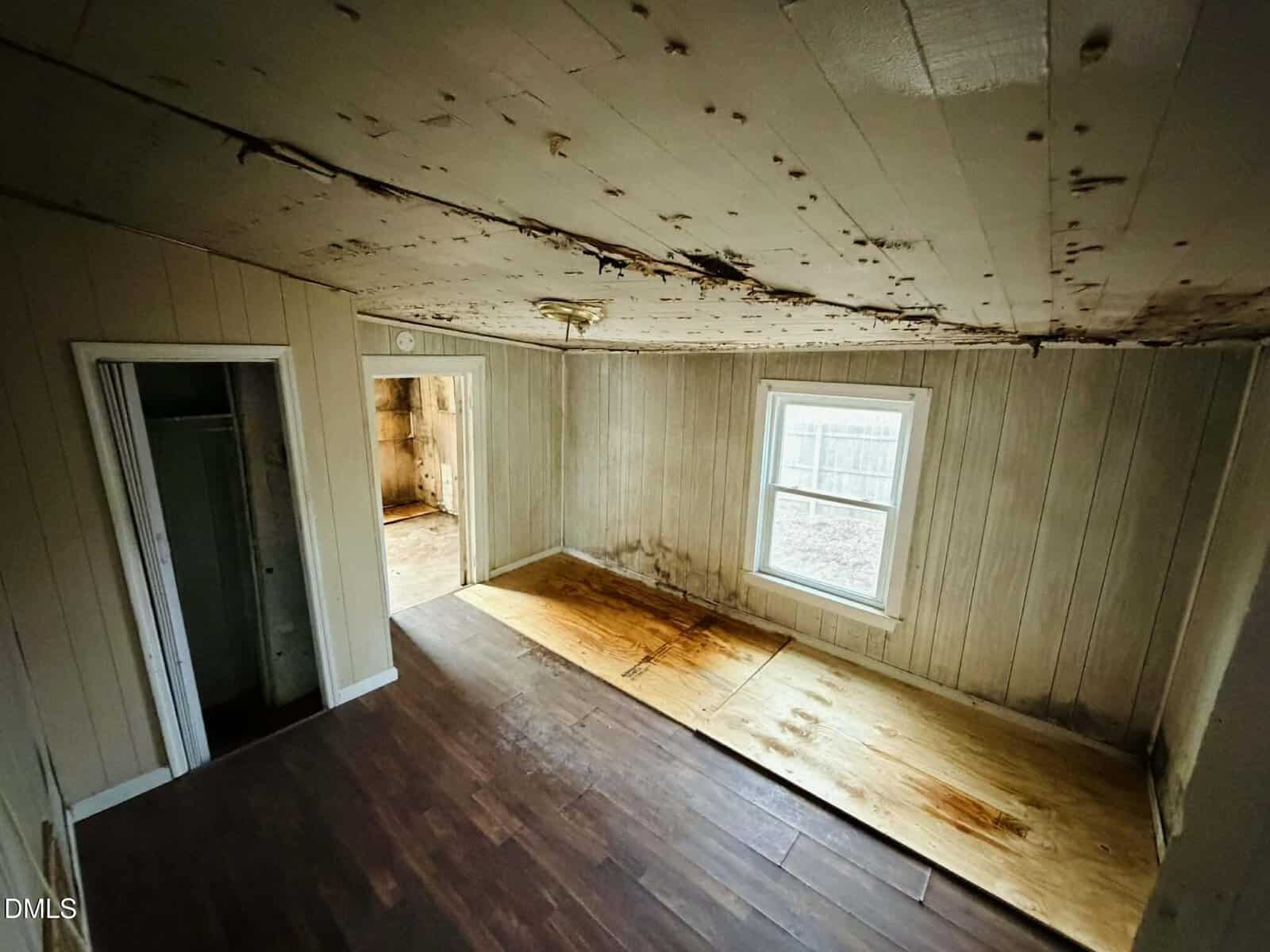 An empty room with peeling walls, water stains, and a damaged ceiling. The floor has dark wood and damaged areas with exposed plywood. Two doors lead to other rooms. Light comes from a single window.