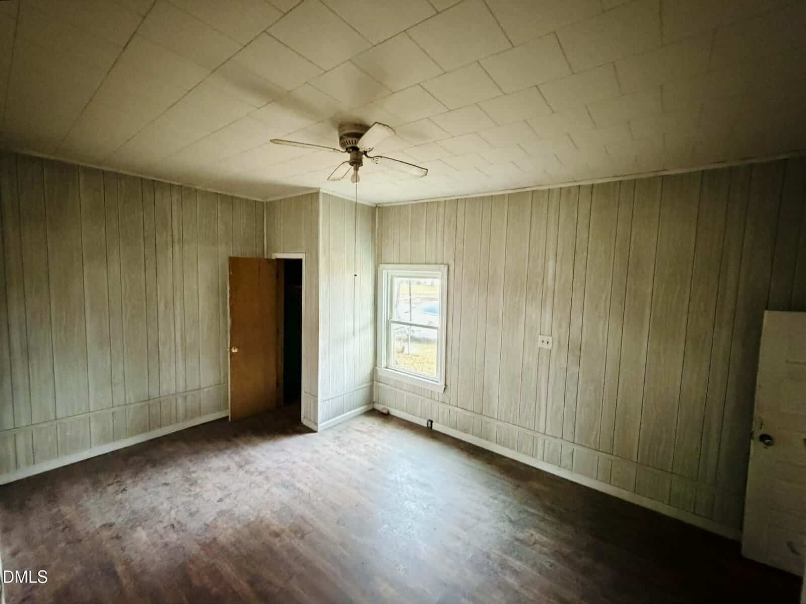 An empty room with wood-paneled walls, a window, an open door, white ceiling tiles, a ceiling fan, and a worn wooden floor. The room appears to be unfurnished and in need of renovation.