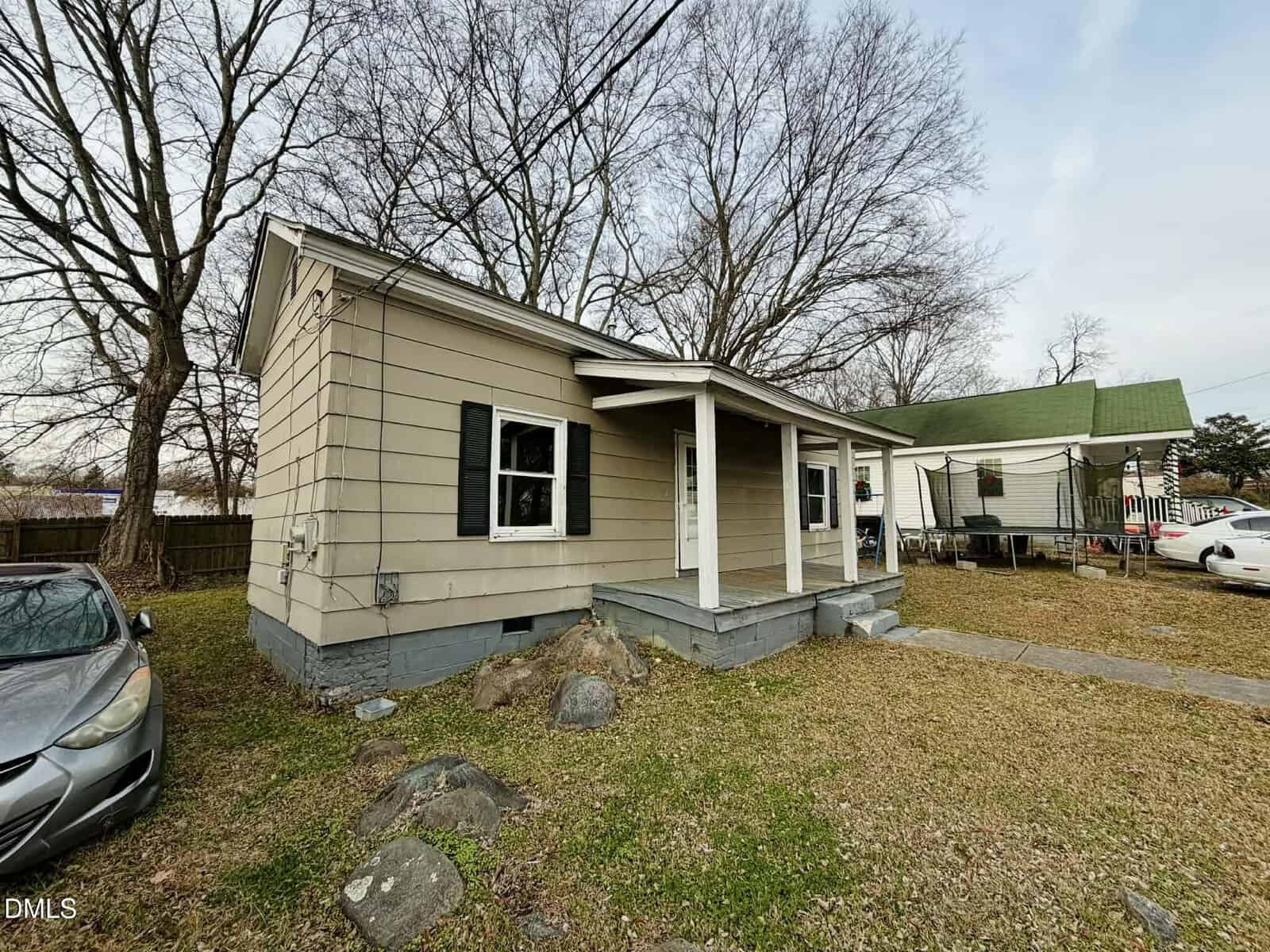 A small, single-story beige house with black shutters and a covered front porch sits on a patchy grass lawn with rocks. A silver car is parked to the left, and leafless trees are in the background.
