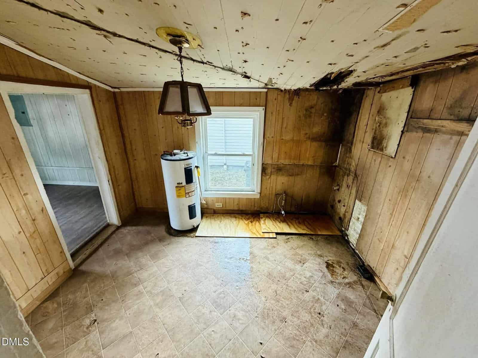 Room with wood-paneled walls, water heater, stained ceiling, and extensive water damage on walls and floor. A single window lets in light; a hanging light fixture is above. Adjacent room with white paneling is visible.