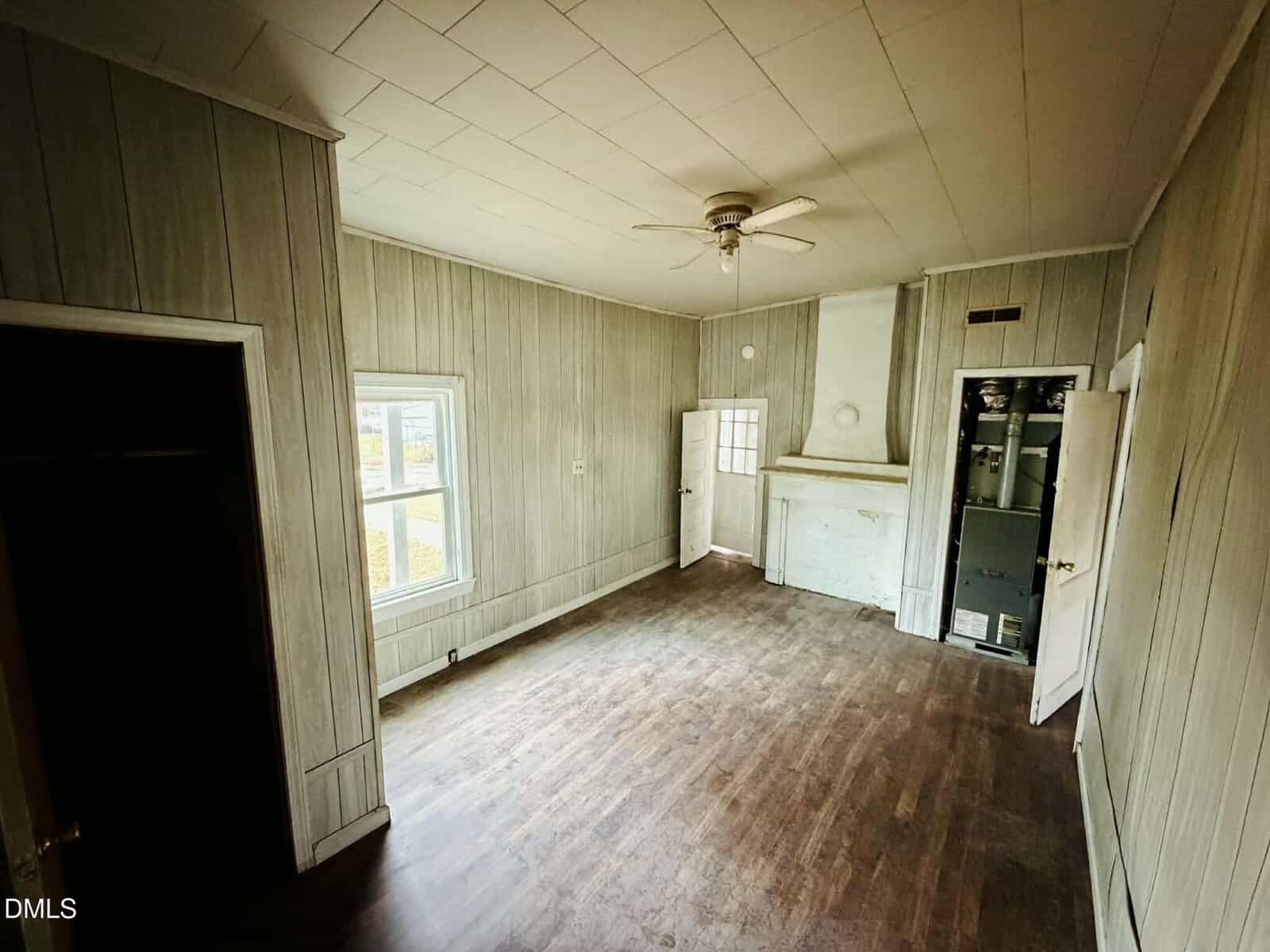A small, empty room with wood-paneled walls, a wood floor, a ceiling fan, two doors, a window, and a wall-mounted cabinet next to a white fireplace. The room appears to be in an older house.