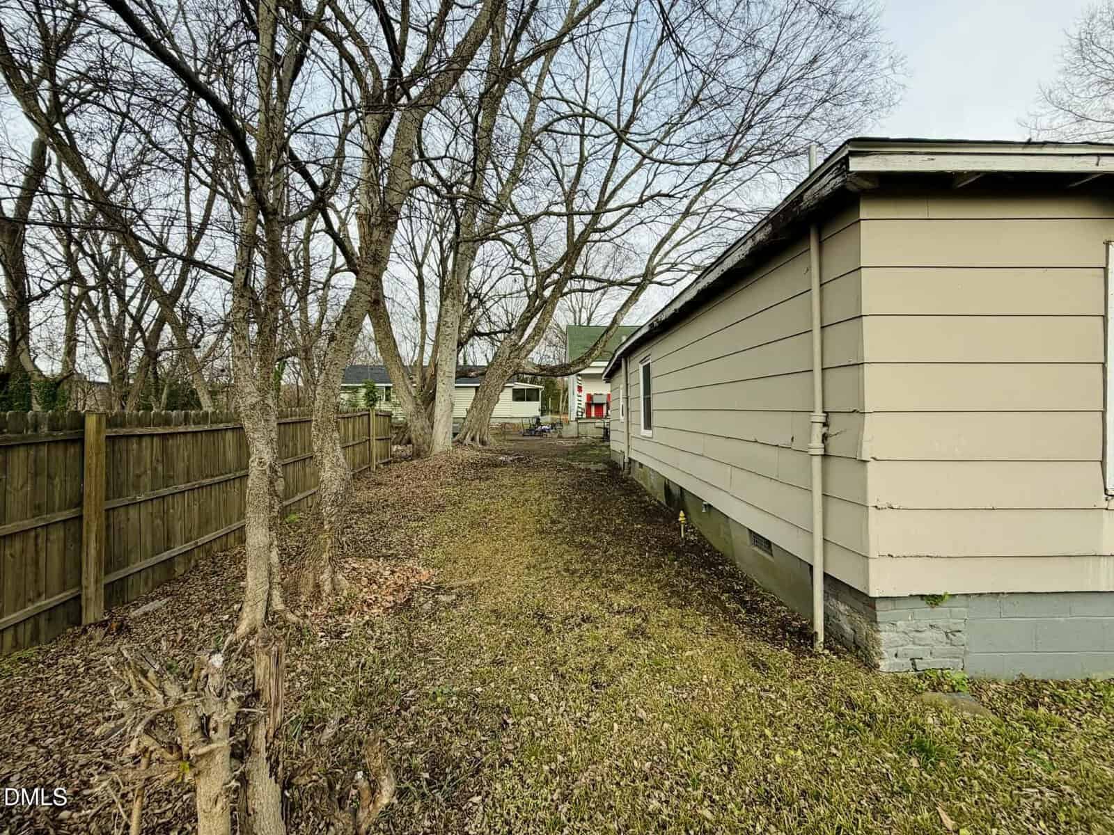 Narrow side yard with patchy grass and fallen leaves, bordered by a wooden fence on the left and a beige house with a gray foundation on the right; leafless trees line the fence.