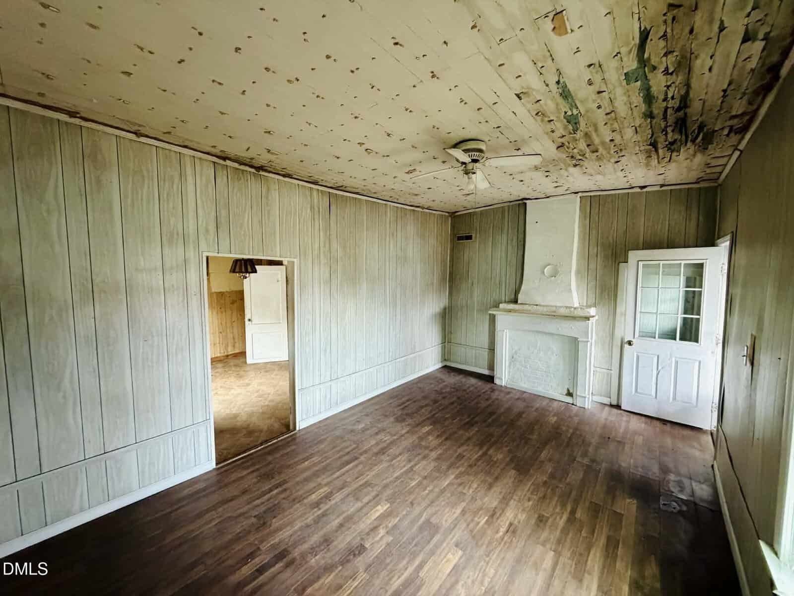 An empty room with wood-style flooring, paneled walls, a white fireplace, and a ceiling fan. The ceiling shows signs of peeling paint and water damage. There are two doorways, one with a glass door and one open.