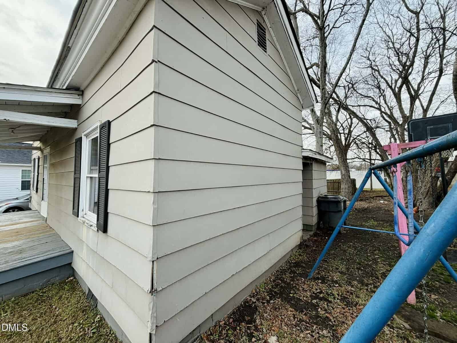 A beige house with horizontal siding and black shutters is shown from the side. There is a small porch, a shed, a trash bin, leaf-covered ground, bare trees, and part of a blue and pink swing set.