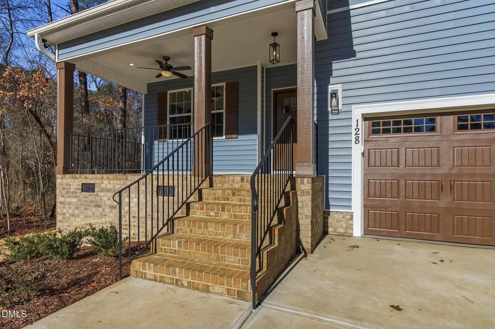 A brick staircase with black railings leads to a covered porch on a blue house with brown shutters and garage door. The porch has columns, a ceiling fan, and a hanging light. House number 128 is visible by the door.