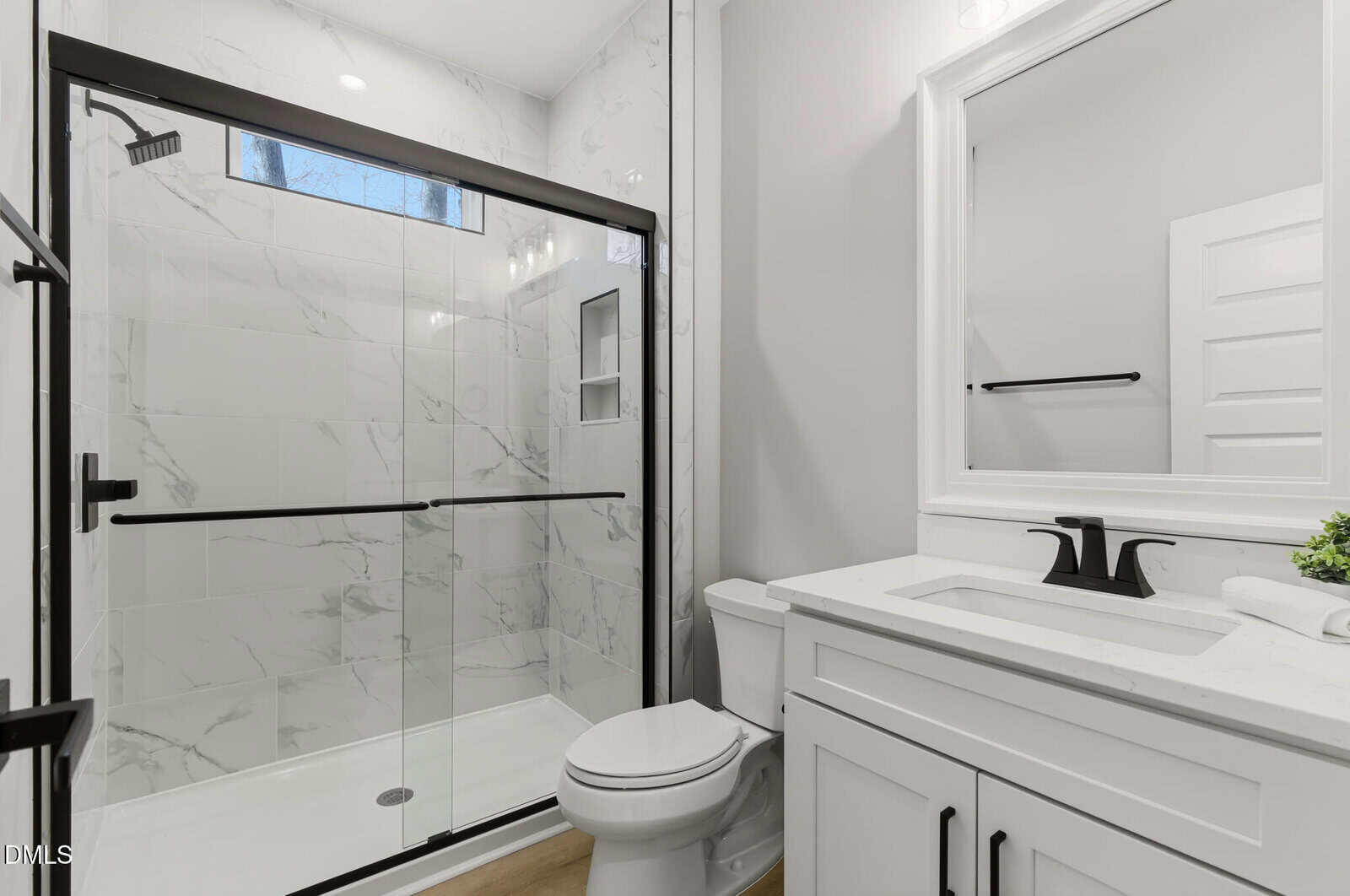Modern bathroom with white cabinets, a white countertop, black fixtures, a toilet, and a glass-enclosed shower with marble-patterned tile. A small plant and rolled towel sit on the counter.