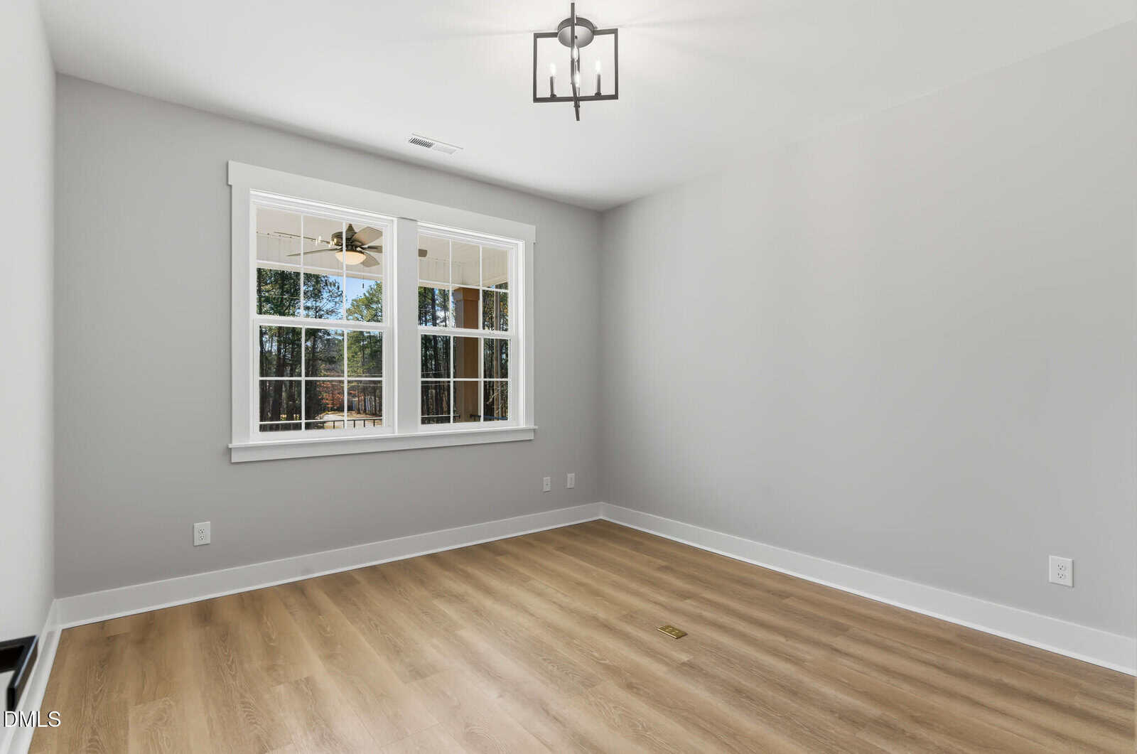 A bright, empty room with light wood flooring, gray walls, white trim, a ceiling light fixture, and a double window showing trees and a ceiling fan outside.