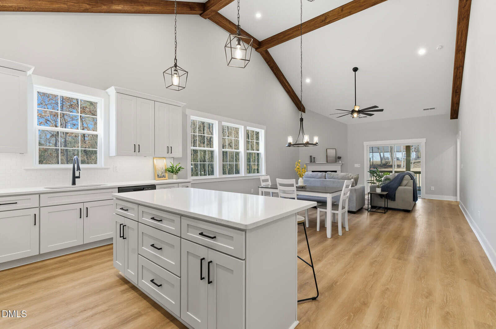 A modern open-concept kitchen and living area with white cabinets, a large island, stainless steel appliances, wood flooring, exposed ceiling beams, pendant lights, and large windows.
