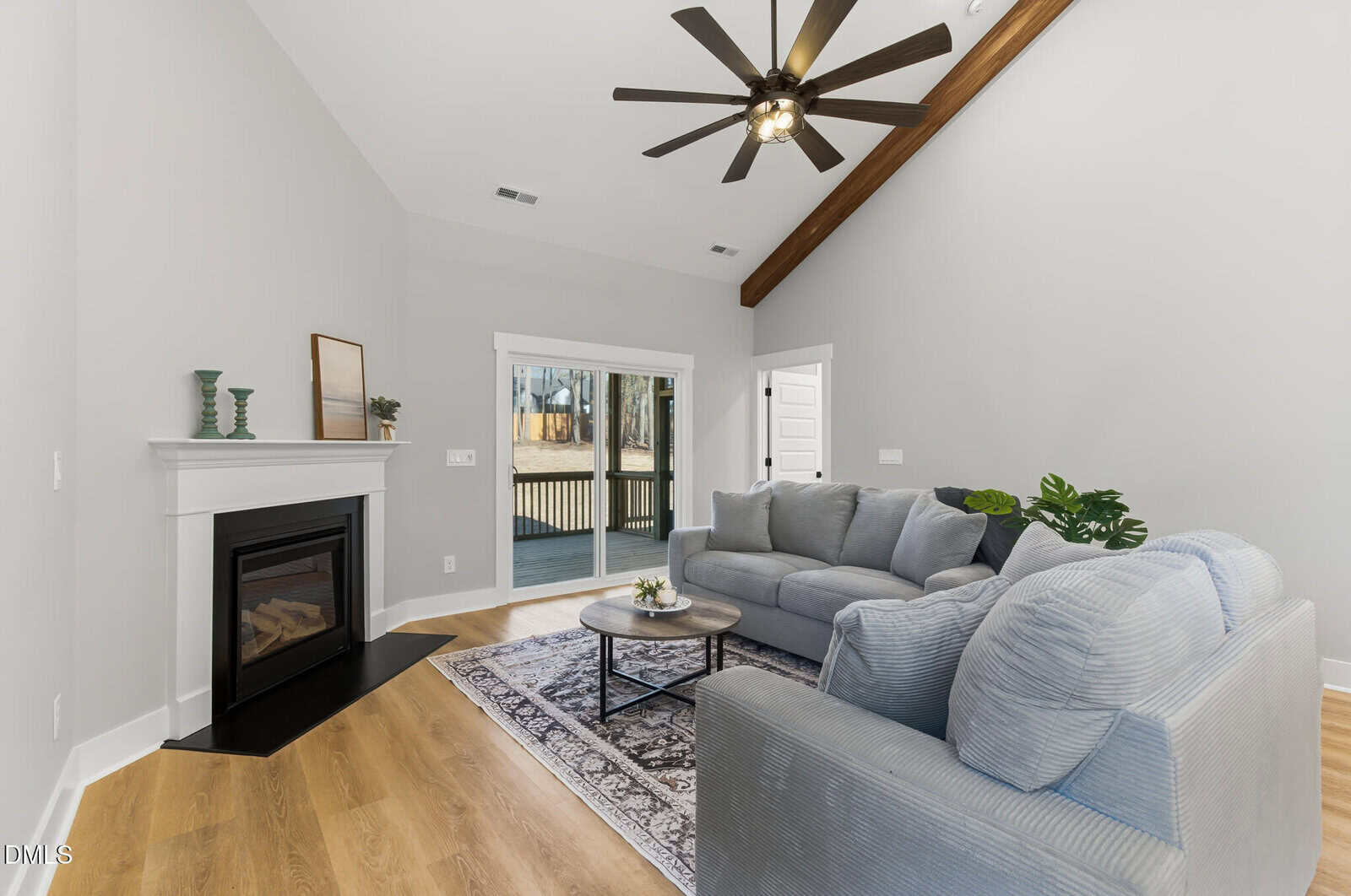 A modern living room with light gray walls, wood flooring, a gray sectional sofa, a round coffee table on a patterned rug, a fireplace with decor, and sliding glass doors leading to a deck.